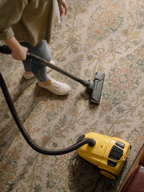 A person using a vacuum cleaner with a yellow canister and black hose to clean a patterned, beige and pastel floral rug in a room, with visible wooden flooring at the edge of the carpet and natural li