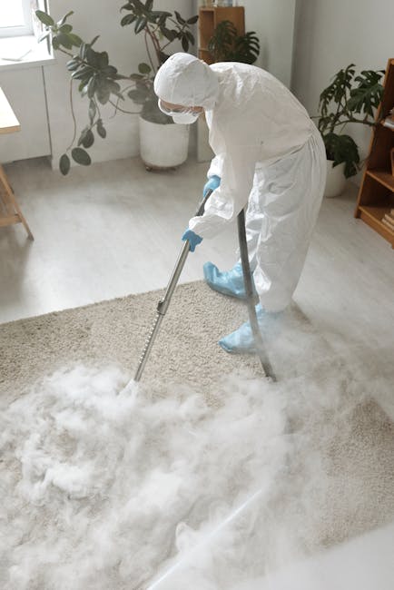 A person using a vacuum cleaner with a yellow canister and black hose to clean a patterned, beige and pastel floral rug in a room, with visible wooden flooring at the edge of the carpet and natural li